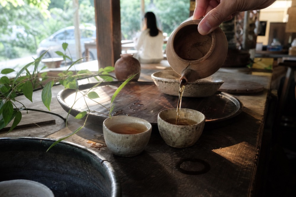Hands pouring amber tea from a small clay pot into cups, no text, no logos, no people
