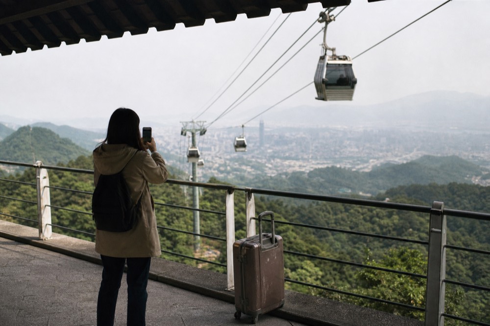 A visitor checking a phone while gondola cabins pass overhead against a cloudy sky, no text, no logos, no people