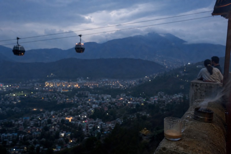 Gondola cabins crossing a valley in low evening light, with the lights of the city just starting to come on below.