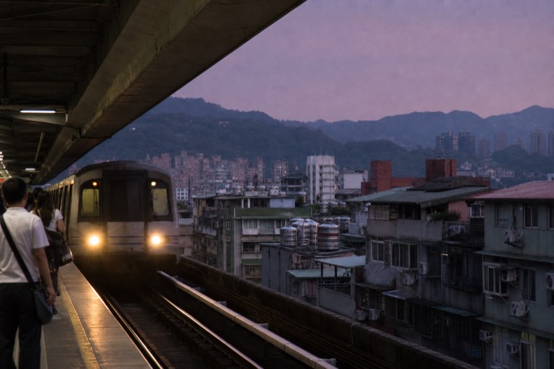 A metro train pulling into an elevated Taipei station platform as the sky dims toward dusk, passengers waiting behind the yellow line.